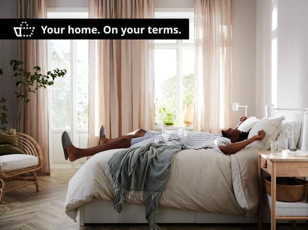 A person laying down on their back on a bed with beige bedding, in a sunny bedrooms with beige and sheer curtains, a black box with white text saying " Your home. On your terms."