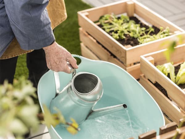 A person pouring water from a can into a large bucket beside several wood planter boxes.