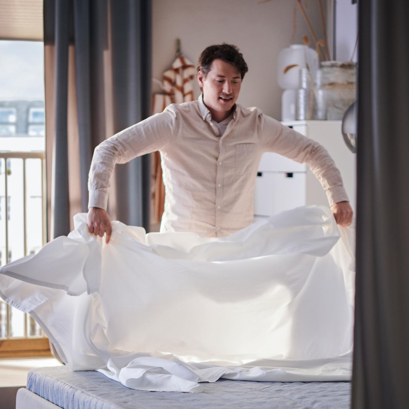 A person puts a sheet on an extra firm, light blue VADSÖ spring mattress on a SLATTUM upholstered bed.