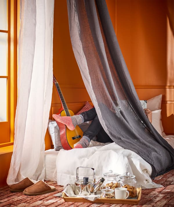 A person relaxing on a spare mattress in the corner of a room behind a canopy of floor length curtains.