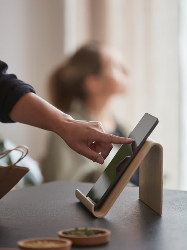 A person scrolls a tablet on a bamboo veneer tablet stand on a black worktop, plus another person sitting by a window.