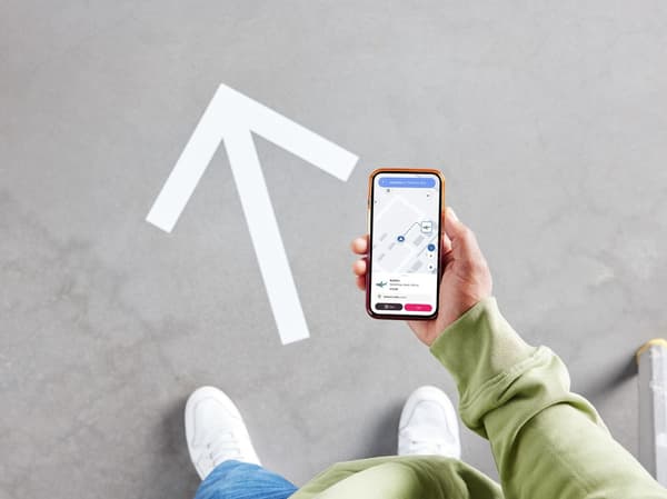 A person seen from above in a store, standing next to a large arrow printed on the floor. They are holding a smartphone displaying a map of the department.