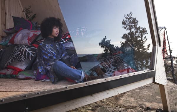 A person sits against a pile of patterned cushions, looking out of an angular window at an ocean view.