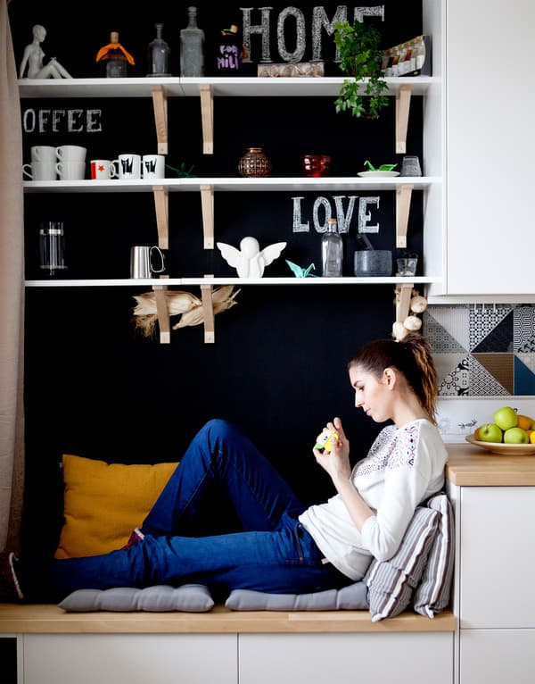 A person sits on a storage bench covered with cushions in this kitchen with white cabinets and a black wall.