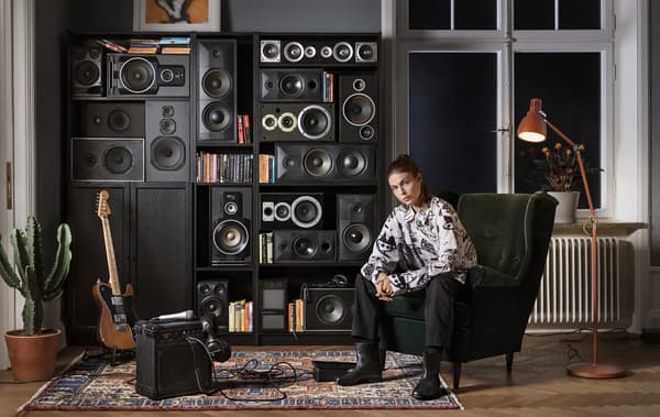 A person sitting in a winged armchair in front of a series of black bookcases filled with speakers.
