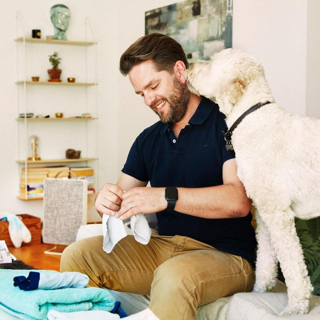 A person sitting on a couch folding laundry while a large white dog stands beside them, nuzzling their ear.