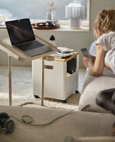 A person sitting on a sofa looking at their phone, with an open laptop on an adjustable side stand and a water bottle in the living room.