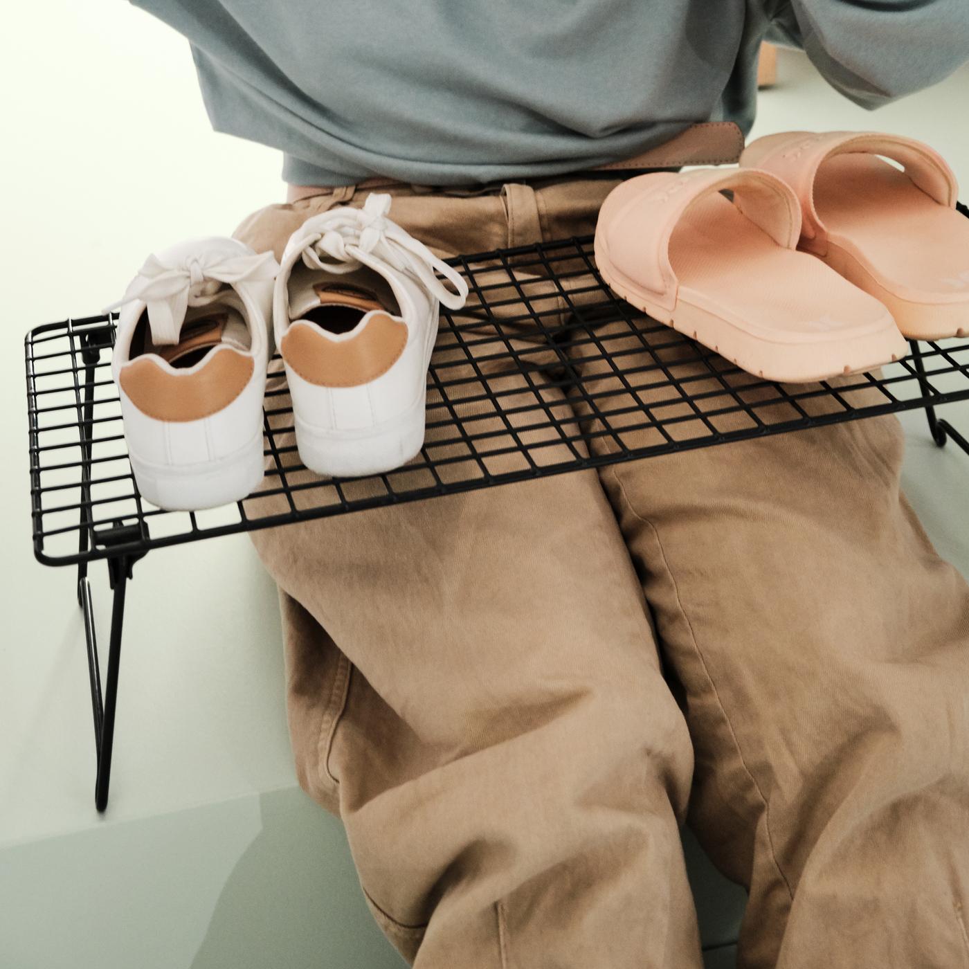 A person sitting on the floor with a grey GREJIG shoe rack over the legs, two pairs of shoes stand on top.