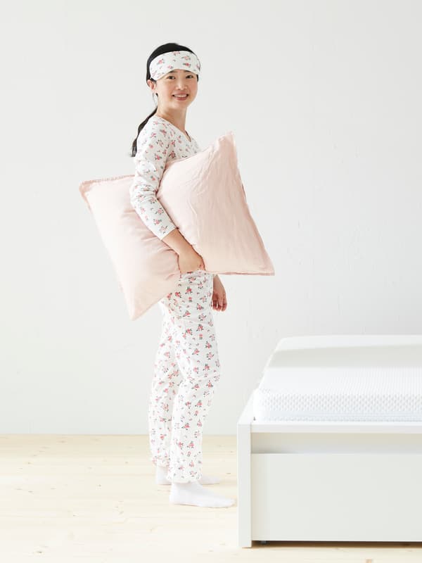 A person smiling wearing PJs and holding a pink pillow in front of a white bed.