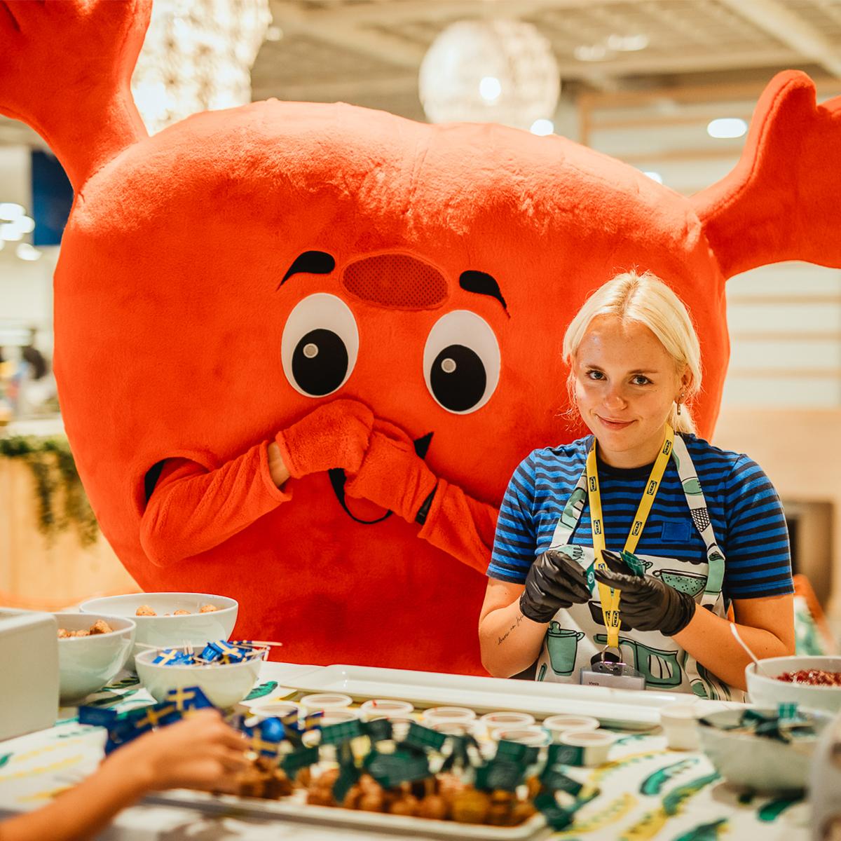 A person standing beside a large red mascot character at a table football game in a brightly lit indoor event space. Several hands are visible playing the game in the foreground, with colorful activity areas and displays in the background.