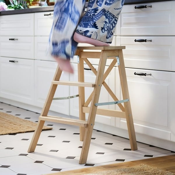 A person stepping up on a BEKVÄM wooden step ladder in a bright kitchen, trying to reach high cabinets.