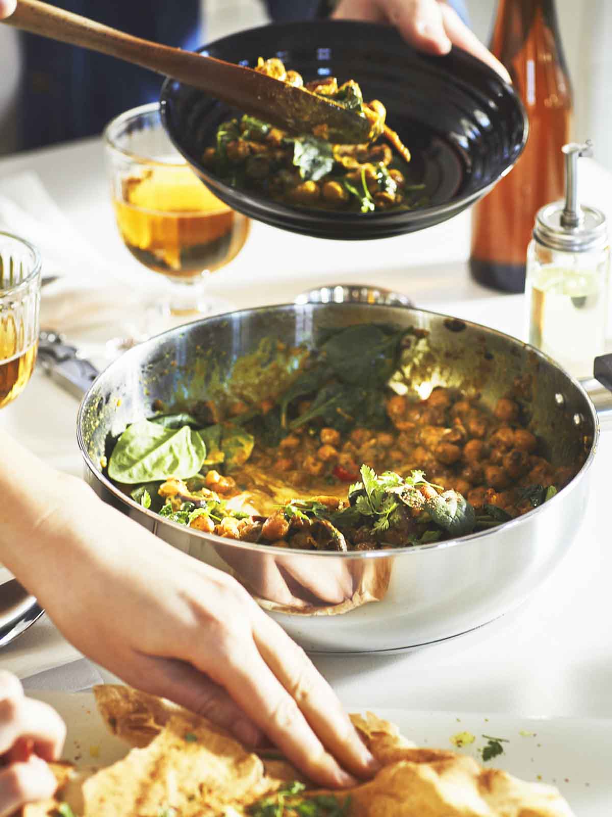 A person uses a wooden spoon to scoop food from a stainless steel MIDDAGSMAT saute pan with lid. 