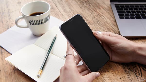A person using a mobile phone, a notebook and coffee cup in front of them and a laptop in the background.
