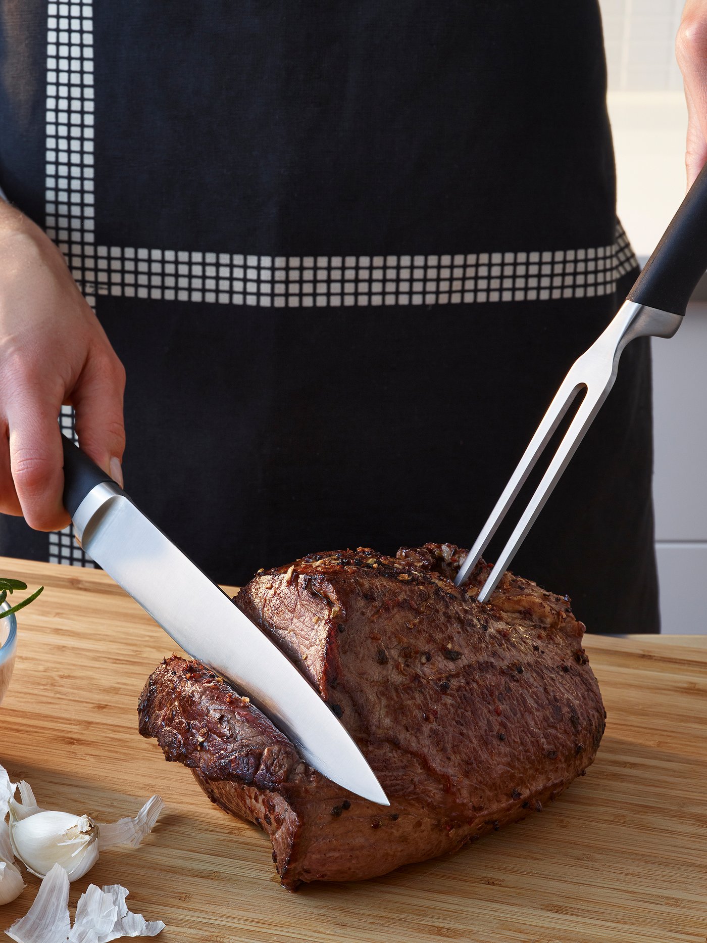 A person wearing a black and white apron is carving meat using a carving knife and fork with black handles.