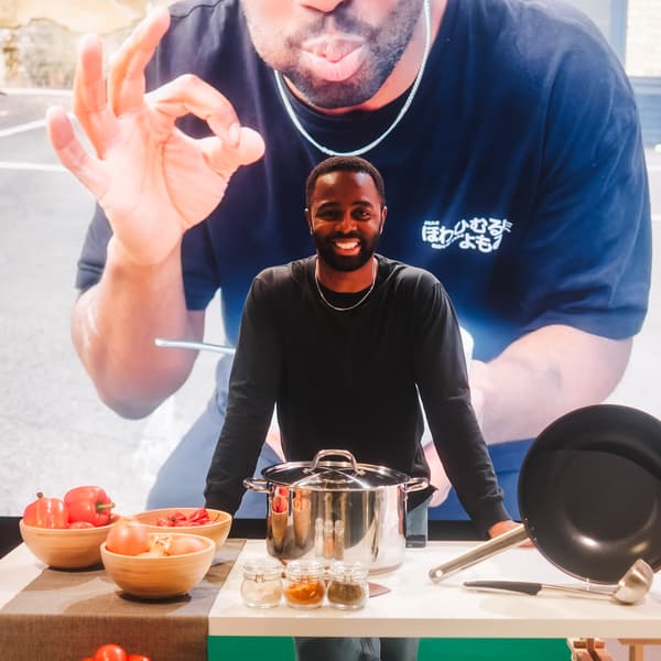 A person wearing a black long-sleeved t-shirt, leaning on a wooden long table set up with cookware, utensils and bowls. A screen in the background shows a person.