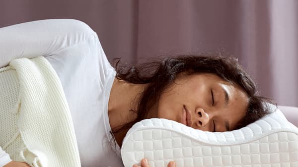 A person wearing a white top asleep on a ROSENSKÄRM pillow on white bed linen with a lilac curtain behind.