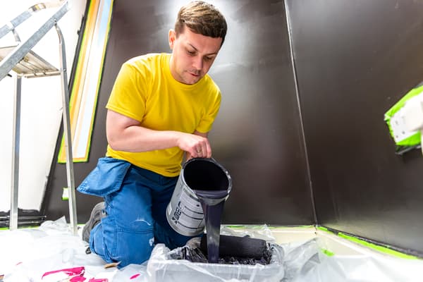 A person wearing a yellow tshirt and blue trousers pours black paint into a tray. Black walls behind.