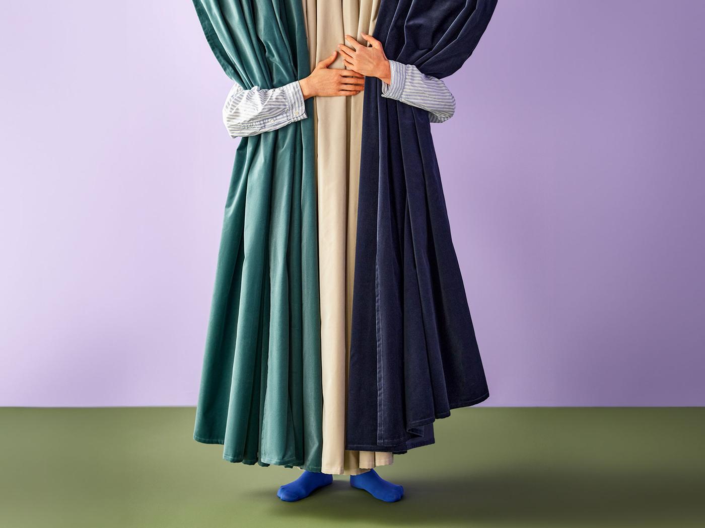 A person with their arms wrapped around three pairs of curtains in green, beige and blue colors.