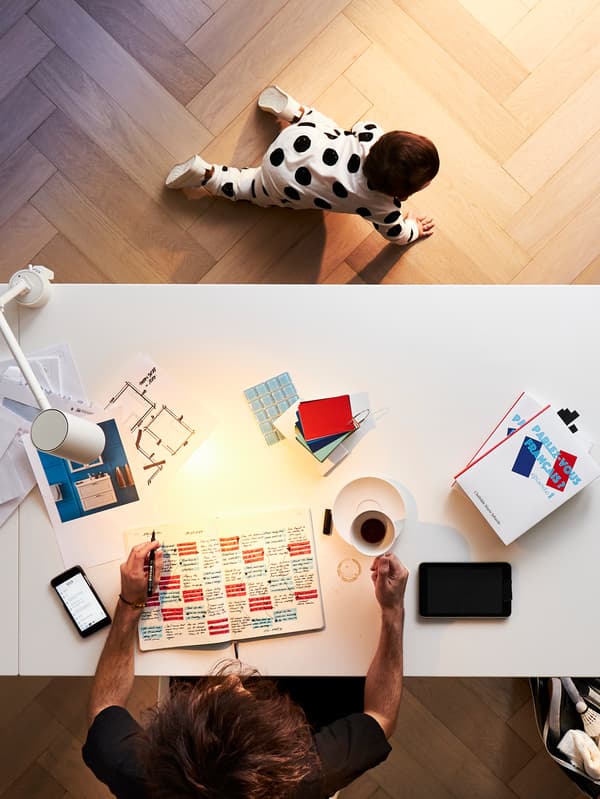 A person working at a home office desk with a cup of coffee and a pen and paper with a baby crawling nearby.