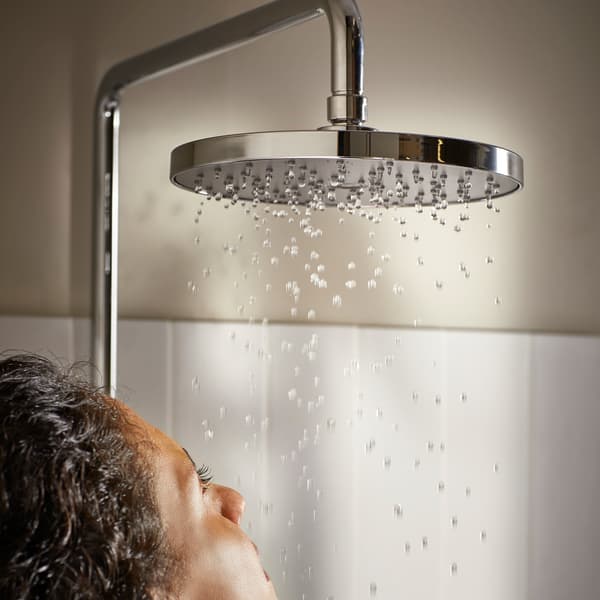 A person’s face is under water droplets that fall from the BROGRUND head shower, in front of a beige wall with white tiles.