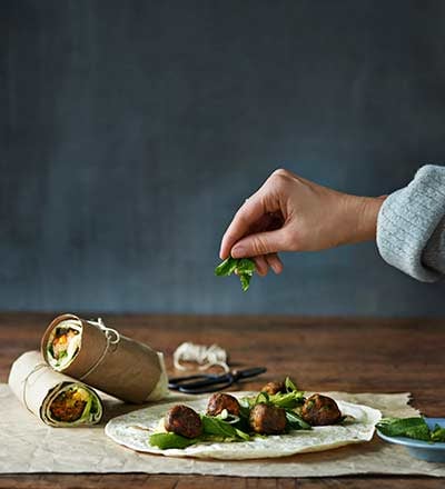 A person’s hand placing fresh herbs onto a tortilla wrap with meatballs and greens, with two wrapped vegetable-filled burritos in the background on a wooden table.