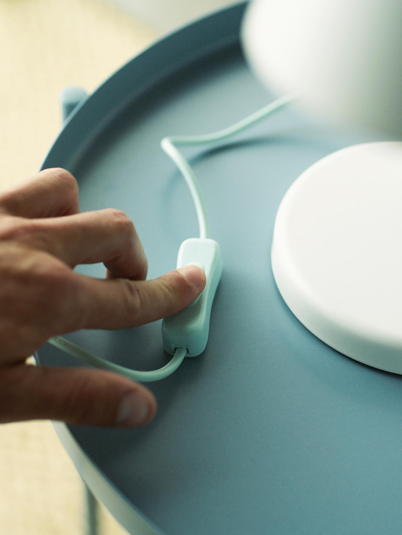A person’s hand presses the on/off switch on the cord of a white FUBBLA LED work lamp on a light-blue tray table.
