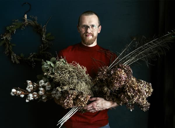 A photo of Łukasz standing in front of a wreath with his arms full of foliage.