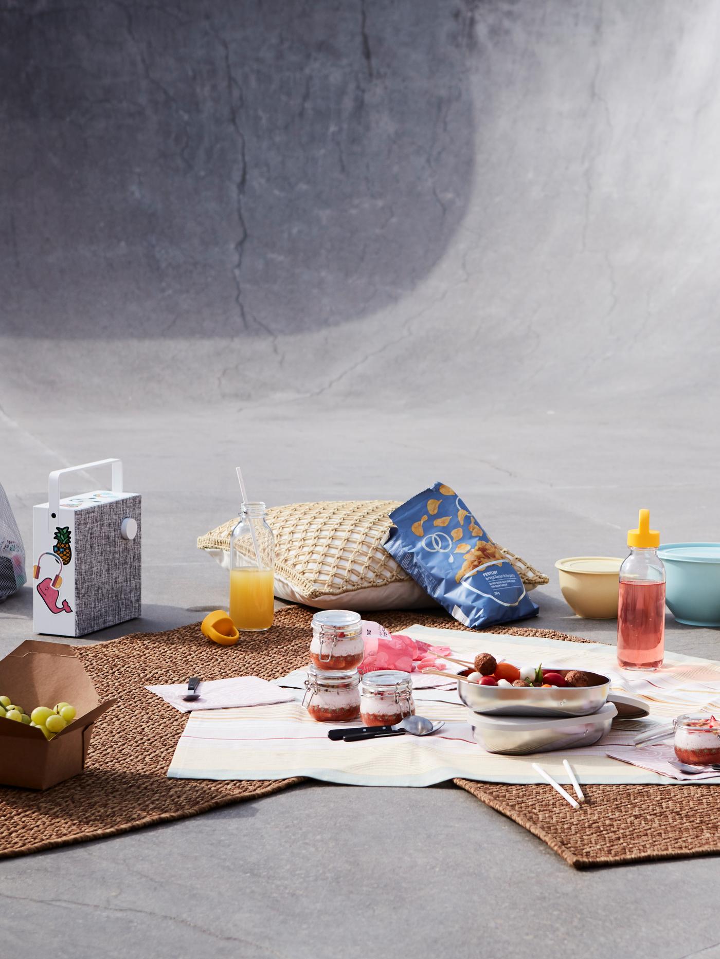 A picnic setting with desserts in glass jars, juice, crisps, an outdoor rug, a speaker and cushions in an urban setting.