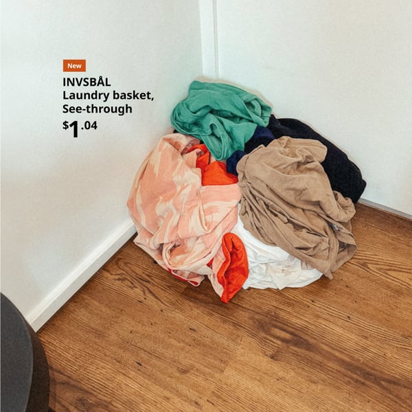 A pile of clothes contained in an INVSBÅL See-through Luandry Basket at the corner of a room with white walls and wooden floors.