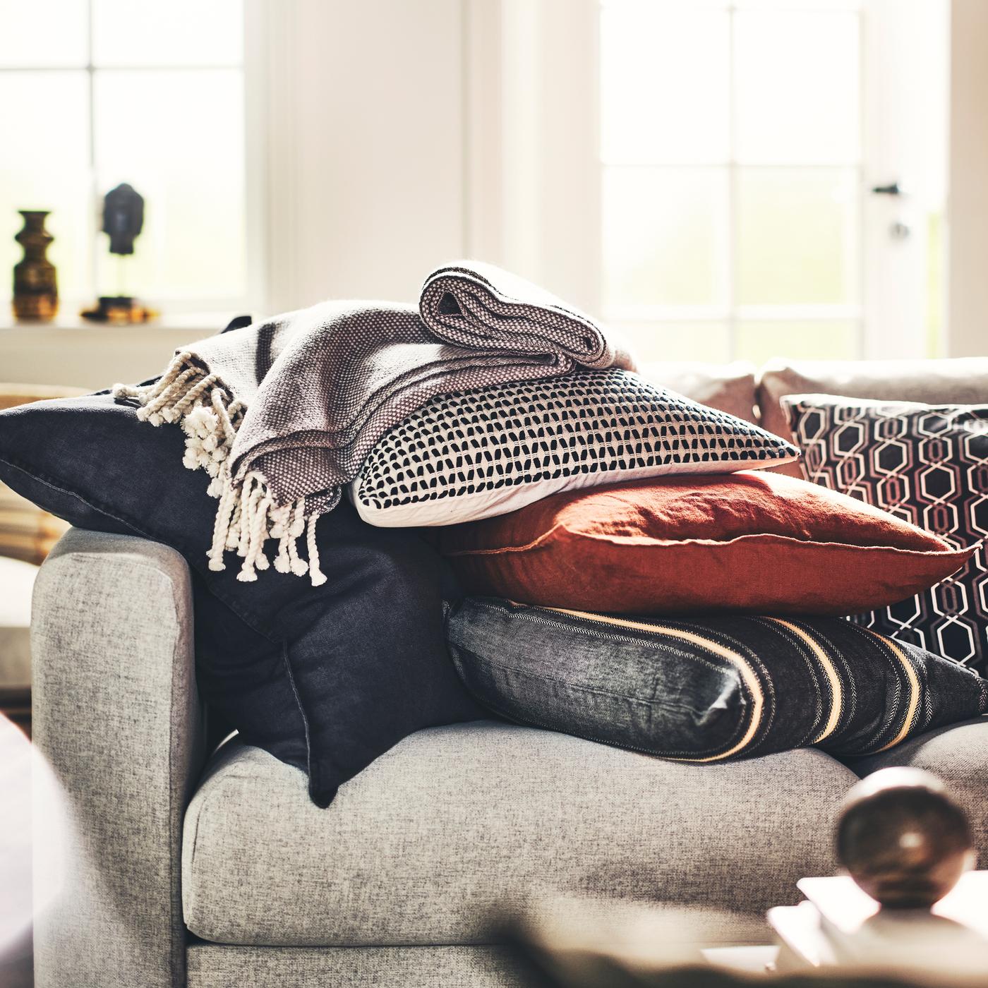 A pile of cushions dressed in a KUSTFLY cushion cover and DYTÅG cushion covers, and a grey/white ULLTISTEL throw, on a sofa.