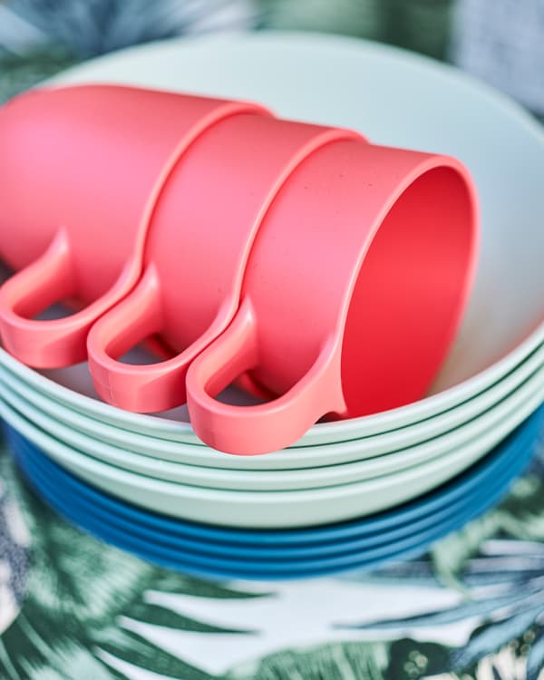 A pile of dark and light blue dishes with three light red mugs stacked on top, on a blue and green leaf print fabric.
