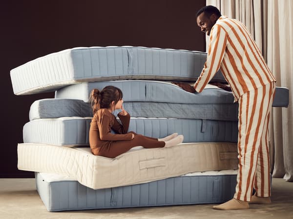 A pile of mattresses with a man in pyjamas pushing down on one mattress and a child sitting on the edge of another.
