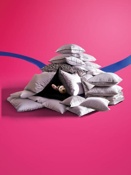 A pillow fort with a FABLER BJÖRN teddy bear soft toy hidding in the middle on a pink background.