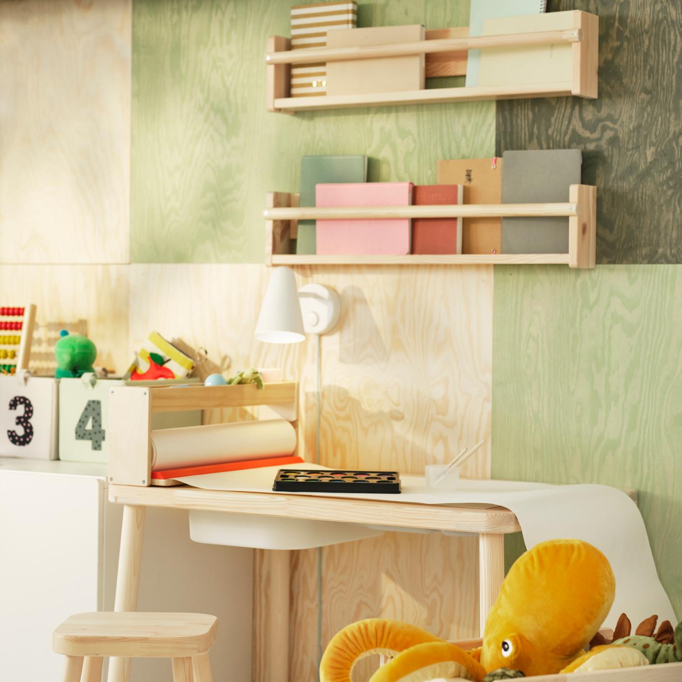 A pine FLISAT children’s table with a TROFAST storage box on top stands under some FLISAT wall storage on the wall.