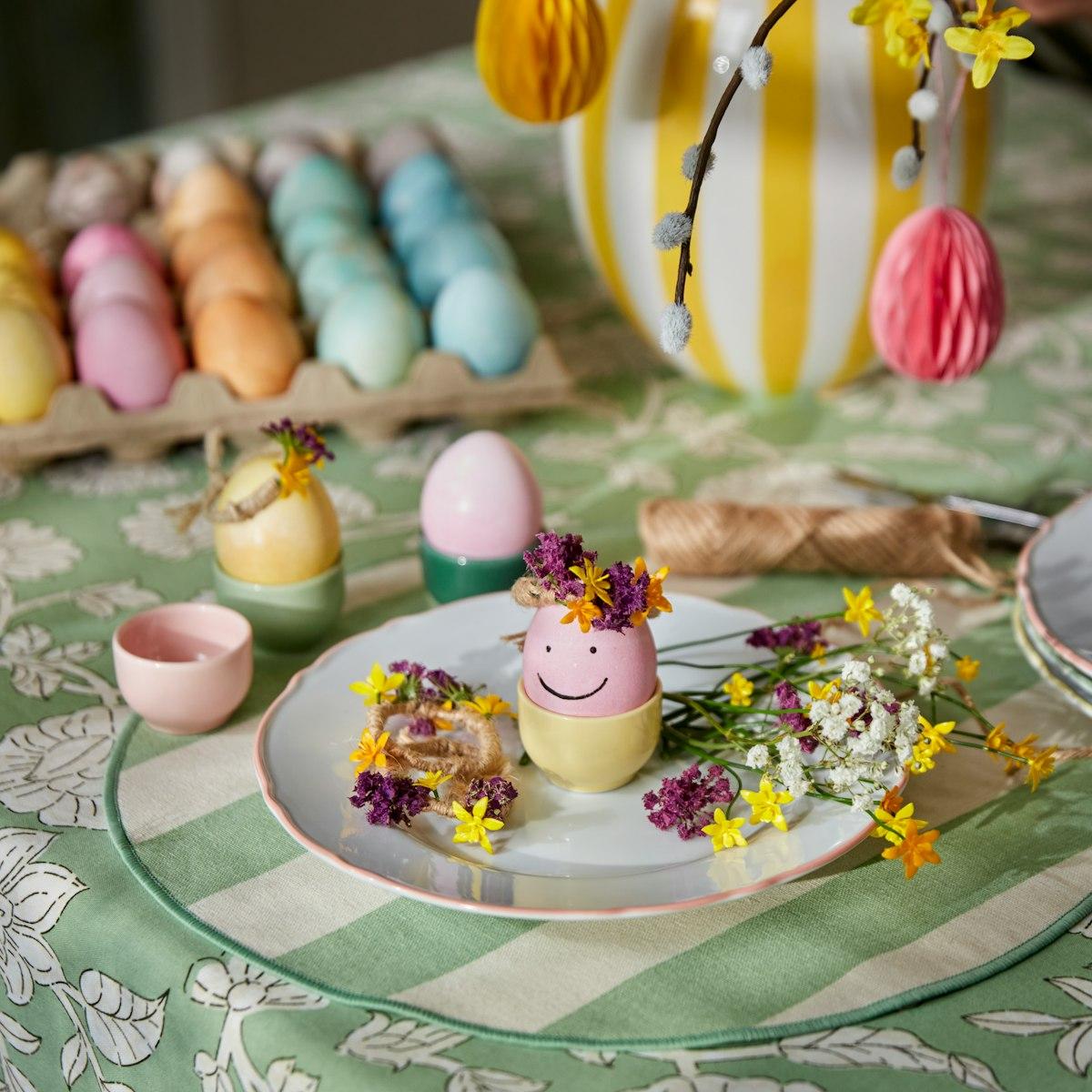 A pink Easter egg with a smiley face and a tiny flower crown sits on a plate on a festive Easter table, surrounded by small colourful flowers and other colourful eggs.
