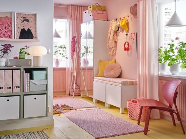 A pink hallway with PLATSA mirrored wardrobe, bench with pink and yellow cushions, a pink rug and hanging hats and scarves.