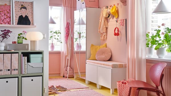 A pink hallway with PLATSA mirrored wardrobe, bench with pink and yellow cushions, a pink rug and hanging hats and scarves.