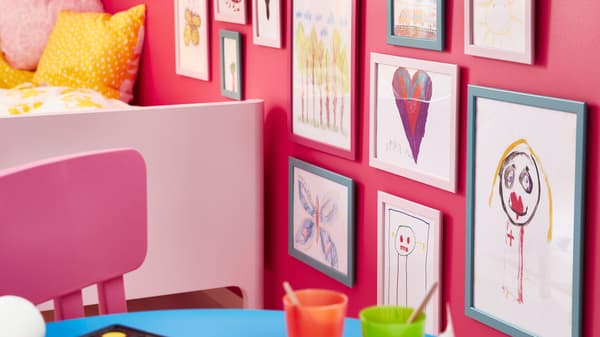 A pink MAMMUT children’s chair sits near a BUSUNGE bed and a wall covered in FISKBO frames holding children’s drawings.