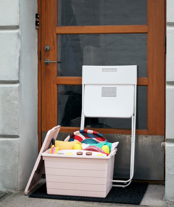 A pink SOCKERBIT box packed for picnic and play on the front step of a building. A folded FEJAN chair leans against the door.