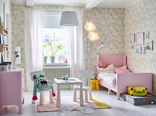 A pink, white and yellow kids' bedroom with extendable BUSUNGE bed in light pink to one side and white MAMMUT tables and chairs in the centre. 