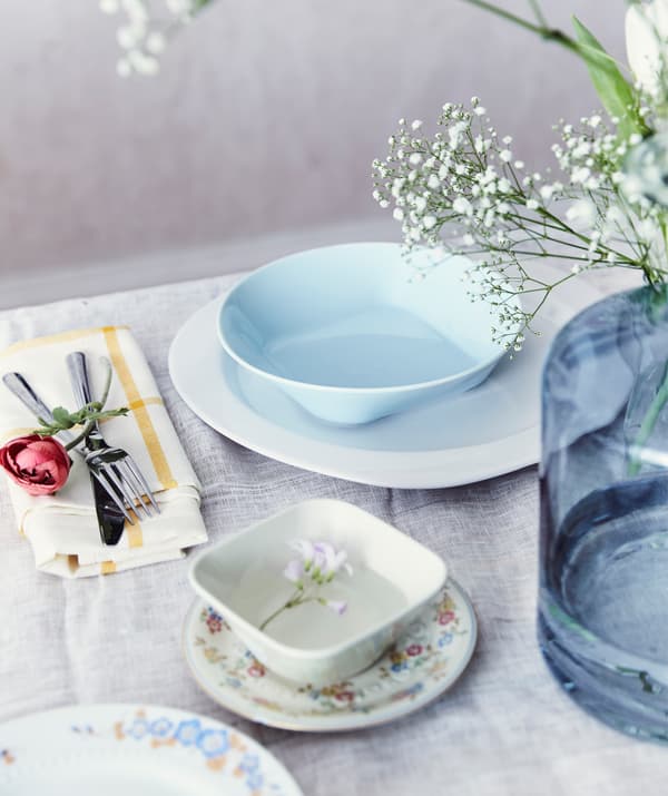 A place setting with a mix of pastel and patterned crockery and a large blue vase of flowers on a white table cloth.