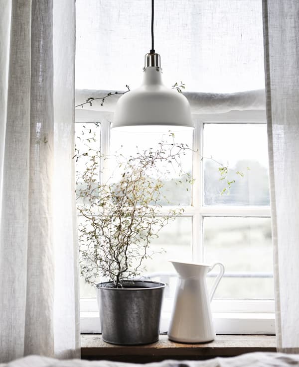 A plant in a dark pot and a white jug on a windowsill with white curtains and a white metal pendant lamp.