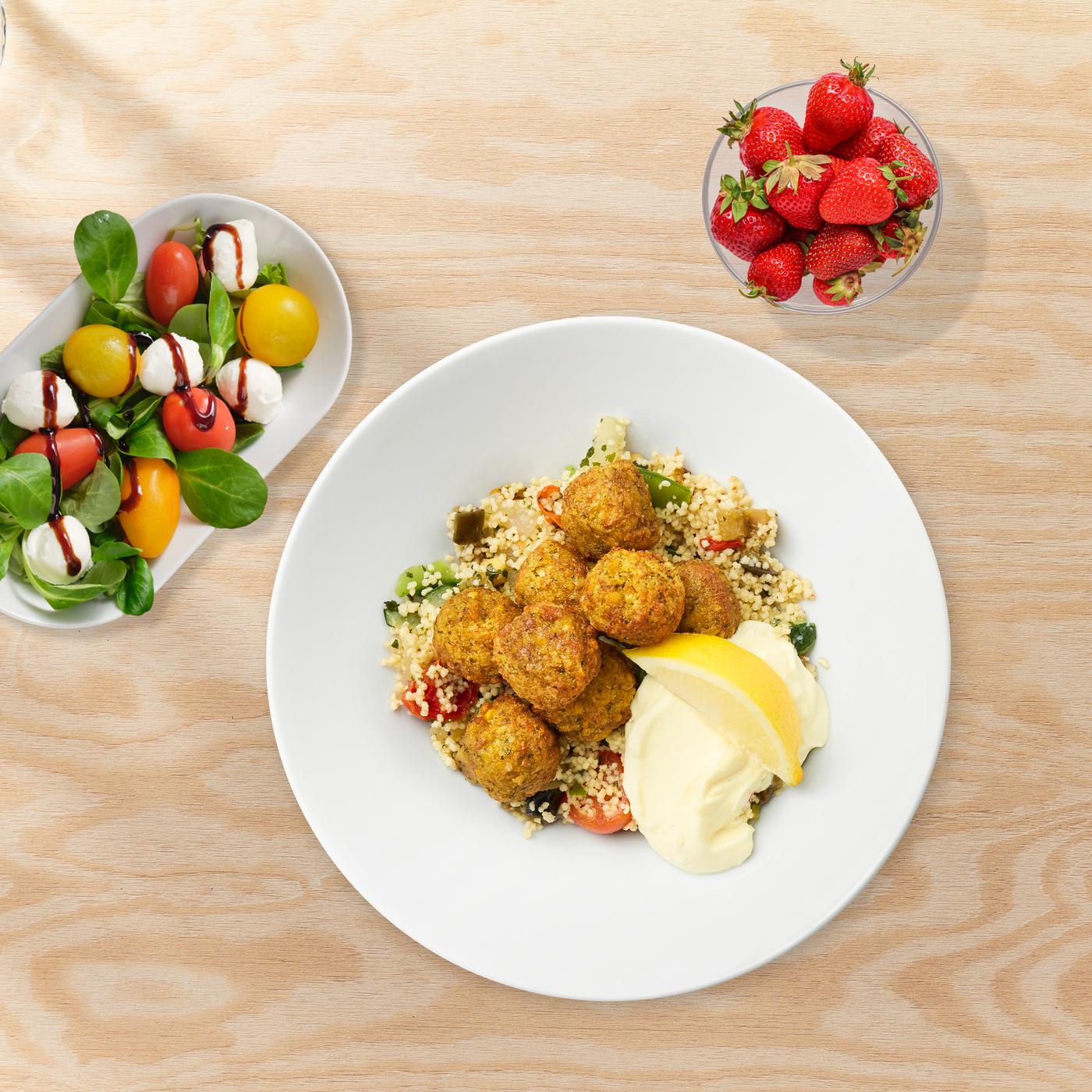 A plate with falafel balls, couscous, aioli, and a lemon wedge, next to a small bowl of tomato-mozzarella salad and a small bowl of fresh strawberries.