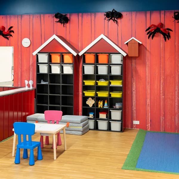 A play area with a small table, colourful chairs and two house-shaped storage shelves filled with boxes, set against a red wooden wall.