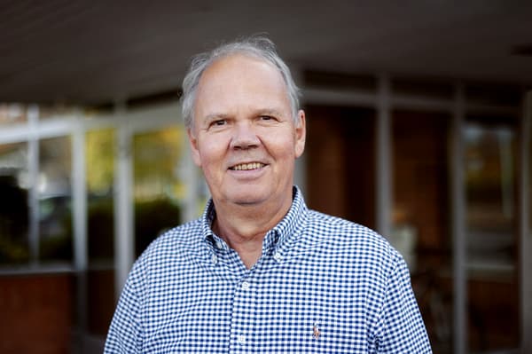 A portrait of an elderly man. He is standing outside and is wearing a small checkered blue shirt.