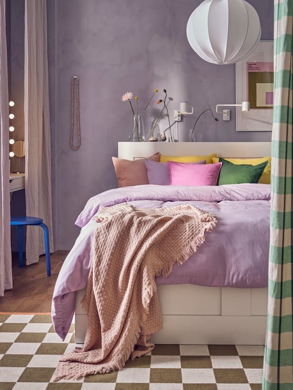 A queen sized BRIMNES bed with storage with lilac NATTSVARMARE duvet quilt cover and bed linen, and cushions in various covers including bright green and pink GURLI covers. A REGNSKUR pendant lamp in white hangs from the ceiling above the bed.