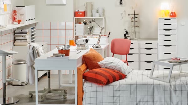 A quirky student room in white and orange furnished with a bed, desk, drawer units, shelves, a bar table and bar stool.