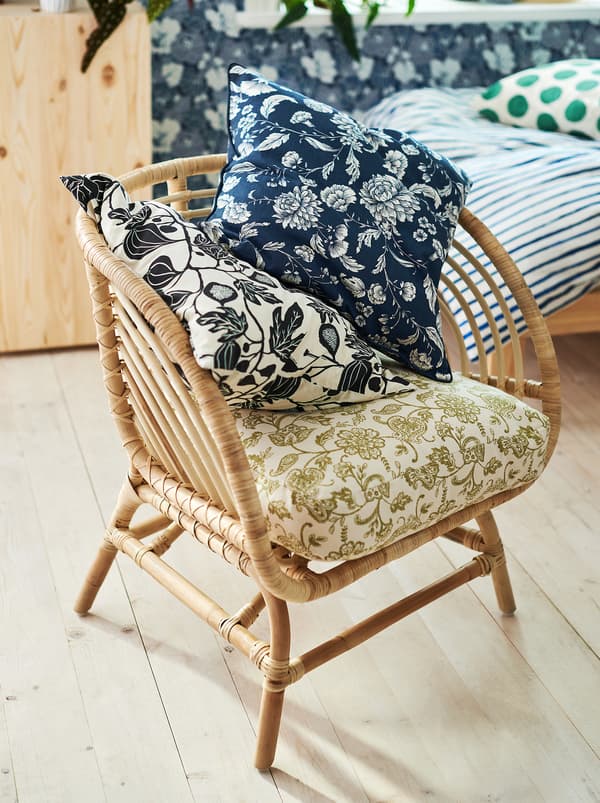 A rattan chair standing in a bedroom with three cushions with slightly different floral patterns in different colours.