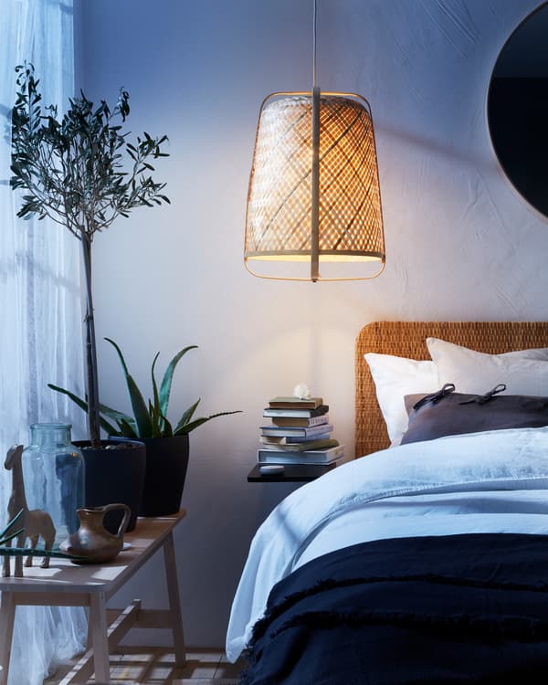 A rattan pendant lamp, a bench, a bed with a rattan headboard, white curtains and different plants in pots.
