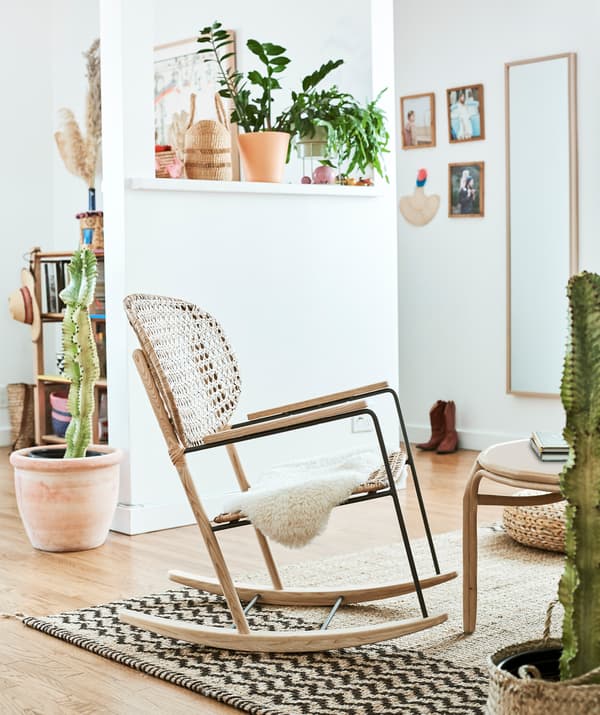 A rattan rocking chair on a hessian rug in a white room, with plants on a shelf and cacti in large pots on the wooden floor.